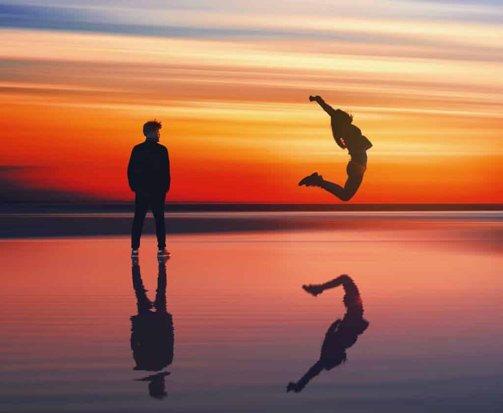 Woman jumping through the air into the water with a beautiful sunset as man watches nearby